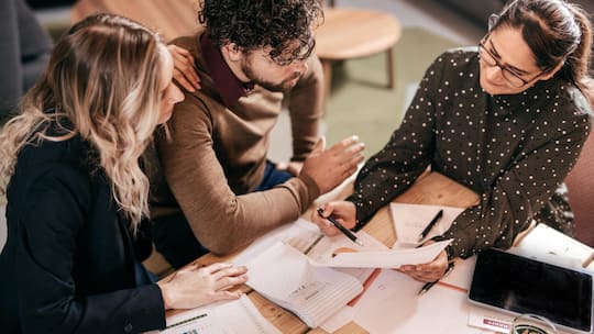 Zwei Frauen und ein Mann diskutieren Finanzdokumente – Drei Millionen Steuerpflichtige in der Schweiz haben kaum oder gar kein Vermögen. Steigende Fixkosten und unerwartete Ereignisse bringen viele Haushalte unter Druck. Wie erleben Sie das? Diskutieren Sie mit.
