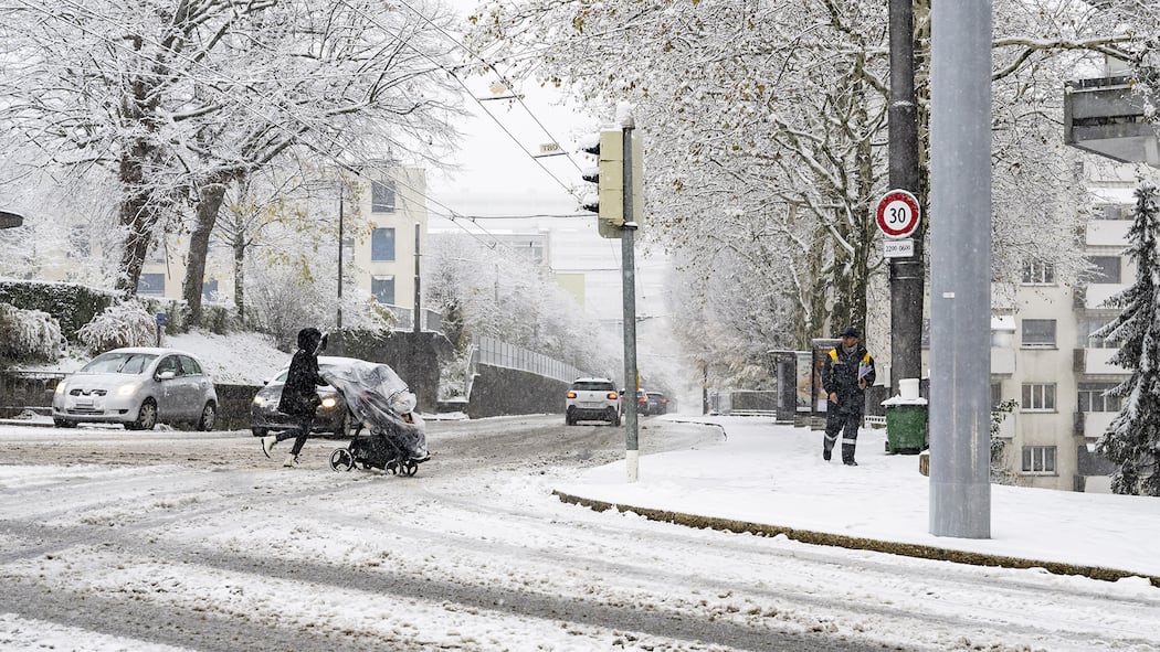 Zu spät im Büro – muss ich länger arbeiten? Eine Person überquert mit einem Kinderwagen die verschneite Strasse in Lausanne. Müssen Arbeitnehmerinnen und Arbeitnehmer länger arbeiten, wenn sie wegen des Schnees und vereister Strassen zu spät zur Arbeit kommen?