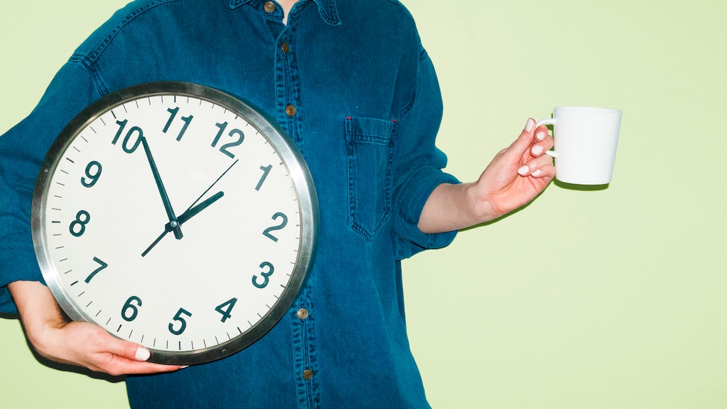 Ausstempeln zum Rauchen? Das muss sein – eigentlich Eine Person mit Kaffeetasse in einer Hand und einer Wanduhr in der anderen Hand vor grünem Hintergrund. Wie Arbeitspausen in der Schweiz geregelt sind, legt das Arbeitsgesetz klar fest.