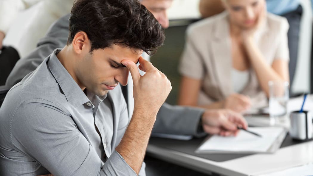 Darf mein Chef die Krankheit erwähnen? Ein Mann stützt seinen Kopf an seine Hand vor einem Tisch mit arbeitenden Personen. Darf der Chef eine Krankheit im Arbeitszeugnis erwähnen?