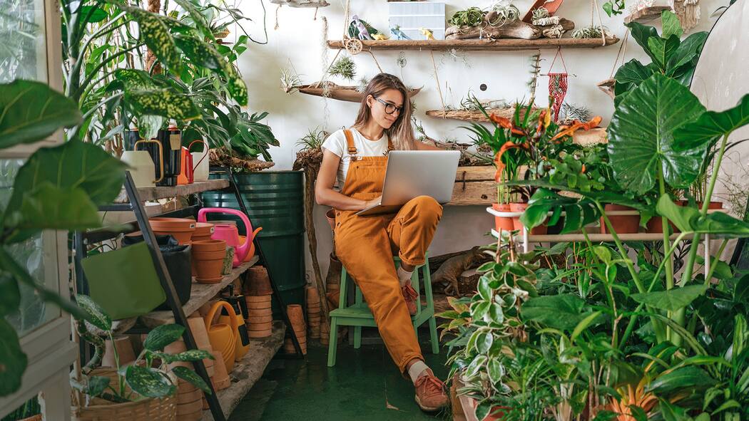 So macht die Krise Firmen innovativ Junge Frau sitzt in einer Gärtnerei auf einem Stuhl und arbeitet am Laptop.