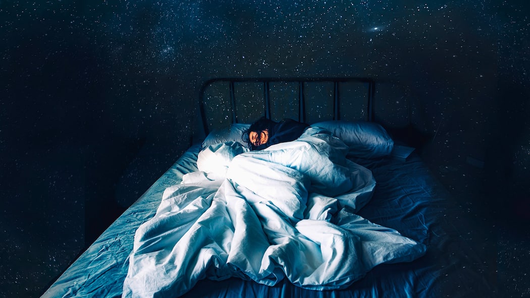 Was für einen gesunden Schlaf förderlich ist Eine Frau schläft tief in ihrem Bett. Im Hintergrund tut sich der Nachthimmel auf.