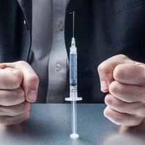 Darf der Chef beim Impfen und Testen mitreden? Ein Mann im Anzug sitzt an einem Tisch, vor ihm steht eine Spritze mit einem Covid-19-Impfstoff. Seine Hände sind zu Fäusten geballt.