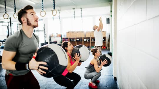 Eine Gruppe junger Menschen steht in einem Fitnessraum und macht Kraftübungen mit schweren Bällen. Um Gewicht zu verlieren, gibt es einige Tipps in Sachen Ernährung. Doch Abnehmen funktioniert nicht ohne sportliche Betätigung.