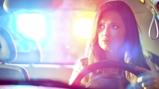 Eine junge Frau sitzt am Steuer während hinter ihr die Blaulichtanlage eines Polizeiautos zu sehen ist. Diese Rechte haben Autolenkerinnen und Autolenker, wenn sie von der Polizei kontrolliert werden