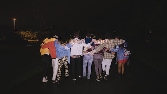 Rear view shot of eight young adults walking down a street at night with their arms round each other.
1125690865