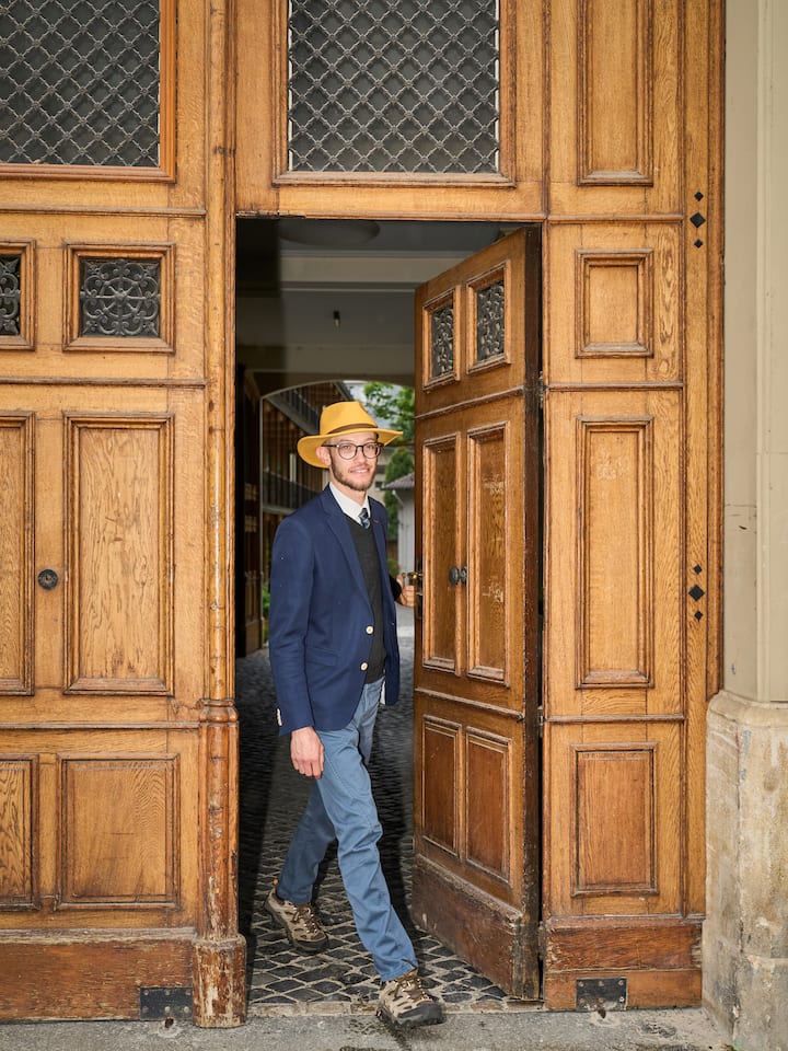 Samuel Zülli tritt lächelnd aus einer Holztüre heraus auf die Strasse, er trägt einen Anzug und einen Hut.
