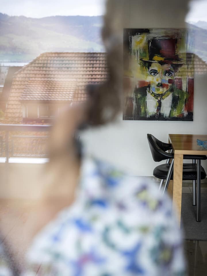 Eine ältere Frau blickt durch ein gespiegeltes Fenster hindurch, im Hintergrund befindet sich ein gemaltes Portrait von Charlie Chaplin - Angehörige von demenzkranken Personen fühlen sich oft einsam, so auch Iris Bürki (Name geändert), deren Ehemann die Diagnose Alzheimer 2021 gestellt bekam. Dem Beobachter erzählt sie ihre Geschichte und wie sie als Angehörige mit der Krankheit und ihrem Mann umgeht.