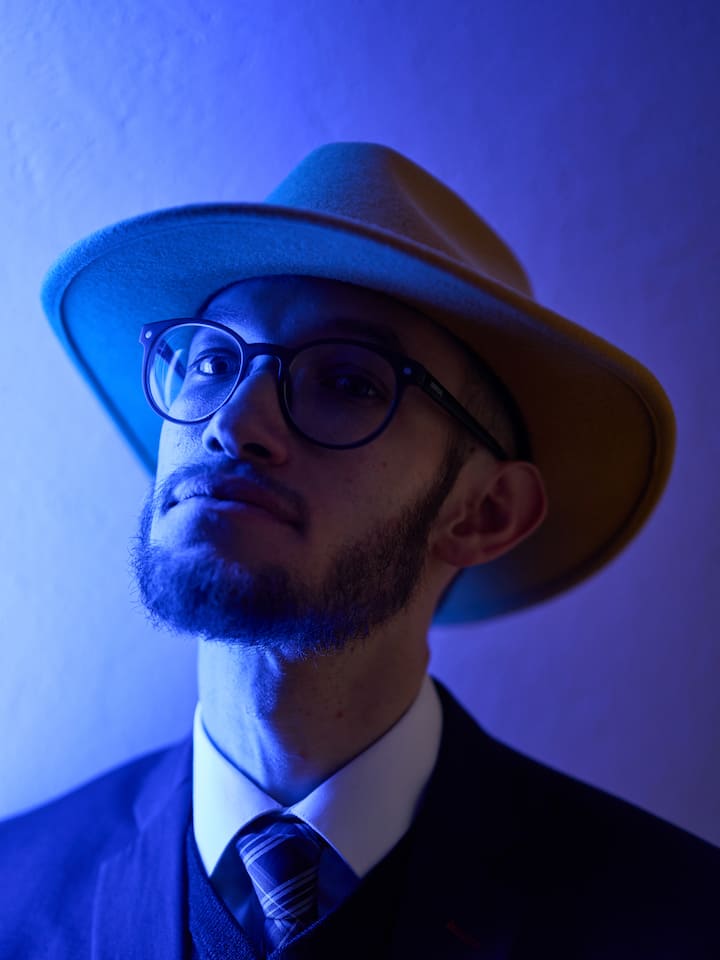 Samuel Zülli blickt ernst in die Kamera, er ist in blaues Licht getaucht und trägt einen Anzug und einen Hut.