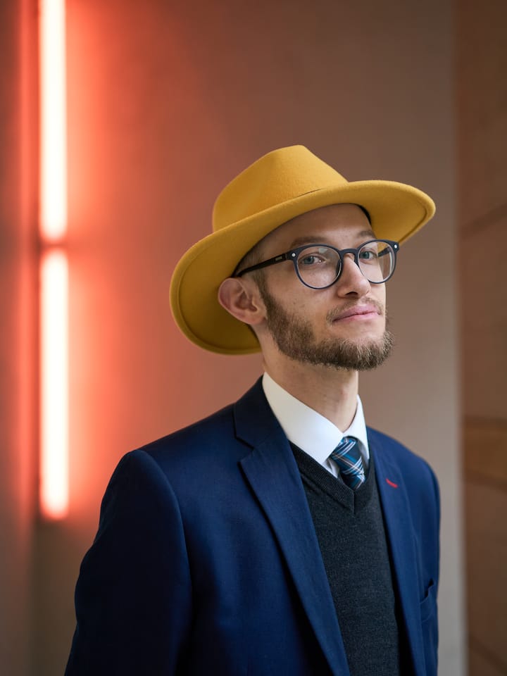 Samuel Zülli steht vor einer rötlichen Wandleuchte, blickt bestimmt in die Kamera, und trägt einen Anzug und einen Hut.