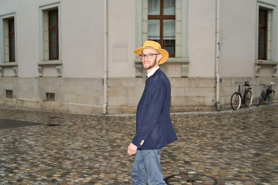 Samuel Zülli geht lächelnd auf einer Strasse, er und trägt einen Anzug und einen Hut.